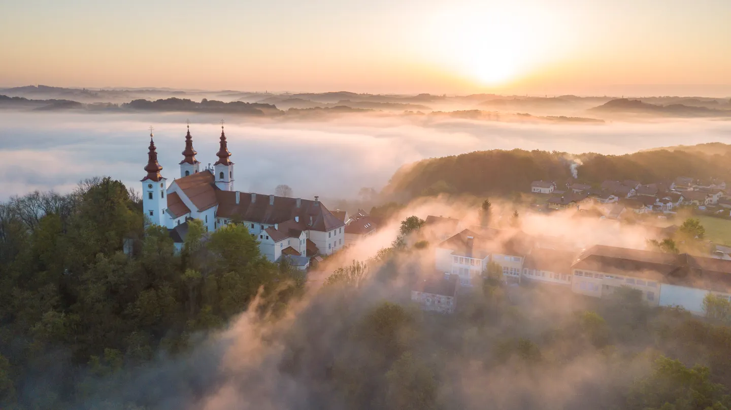 A church and town emerging from warm morning fog beneath the rising sun.