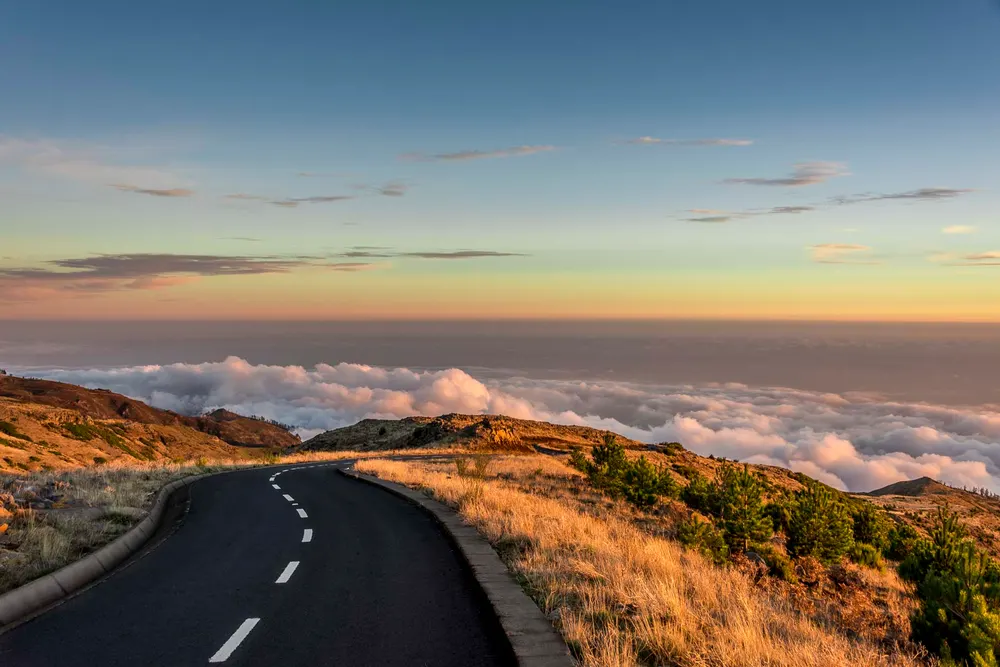 A mountain road curves above a blanket of clouds in warm sunset light.